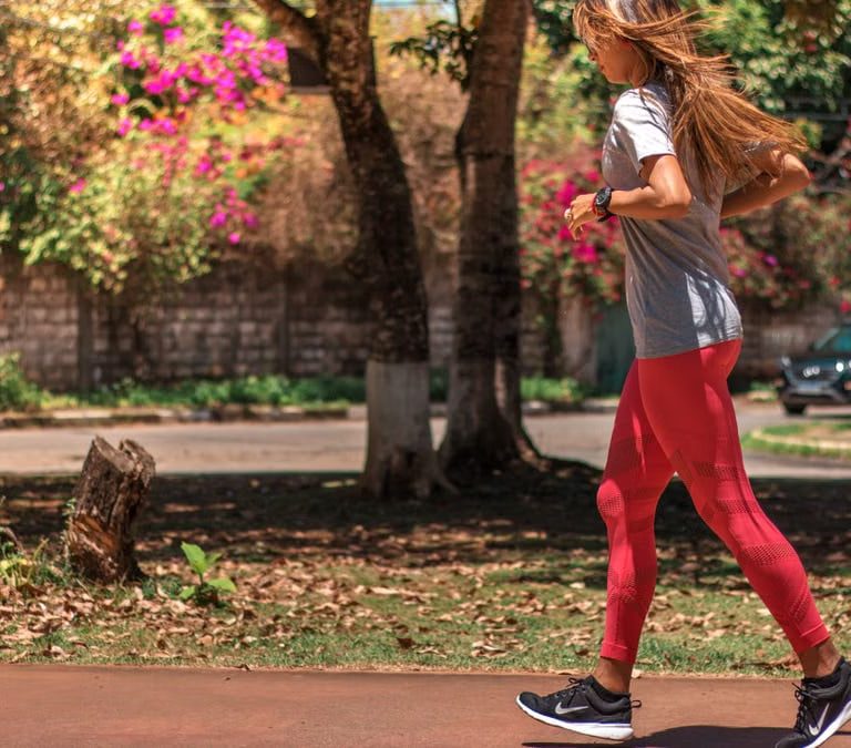 A woman jogs on a path surrounded by trees and vibrant flowers, embodying a healthy lifestyle.